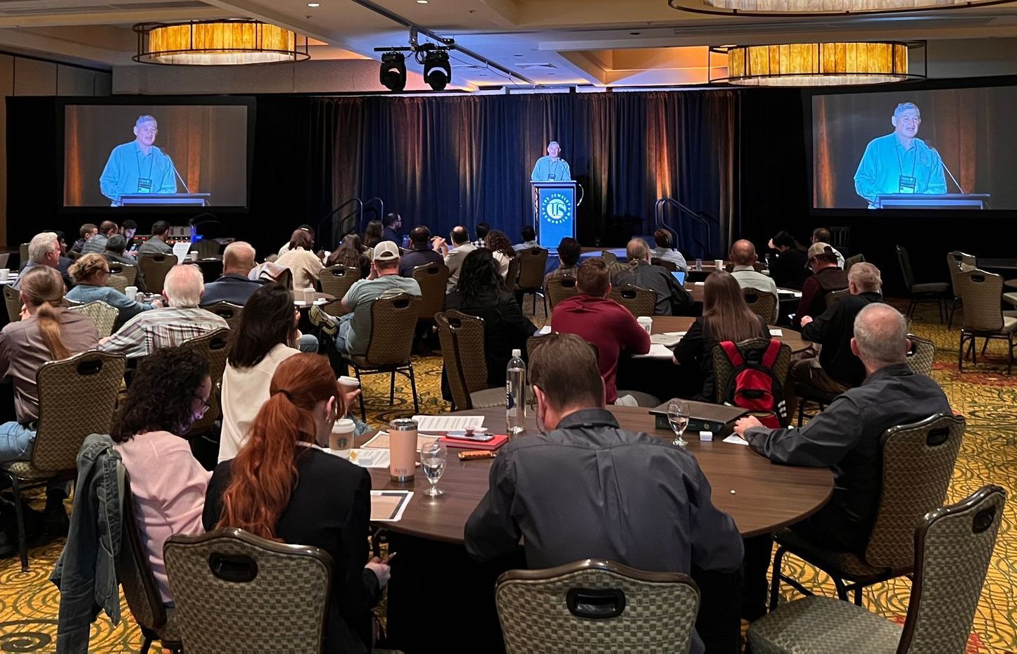Audience at conference listens to speaker on stage with screens showing speaker.