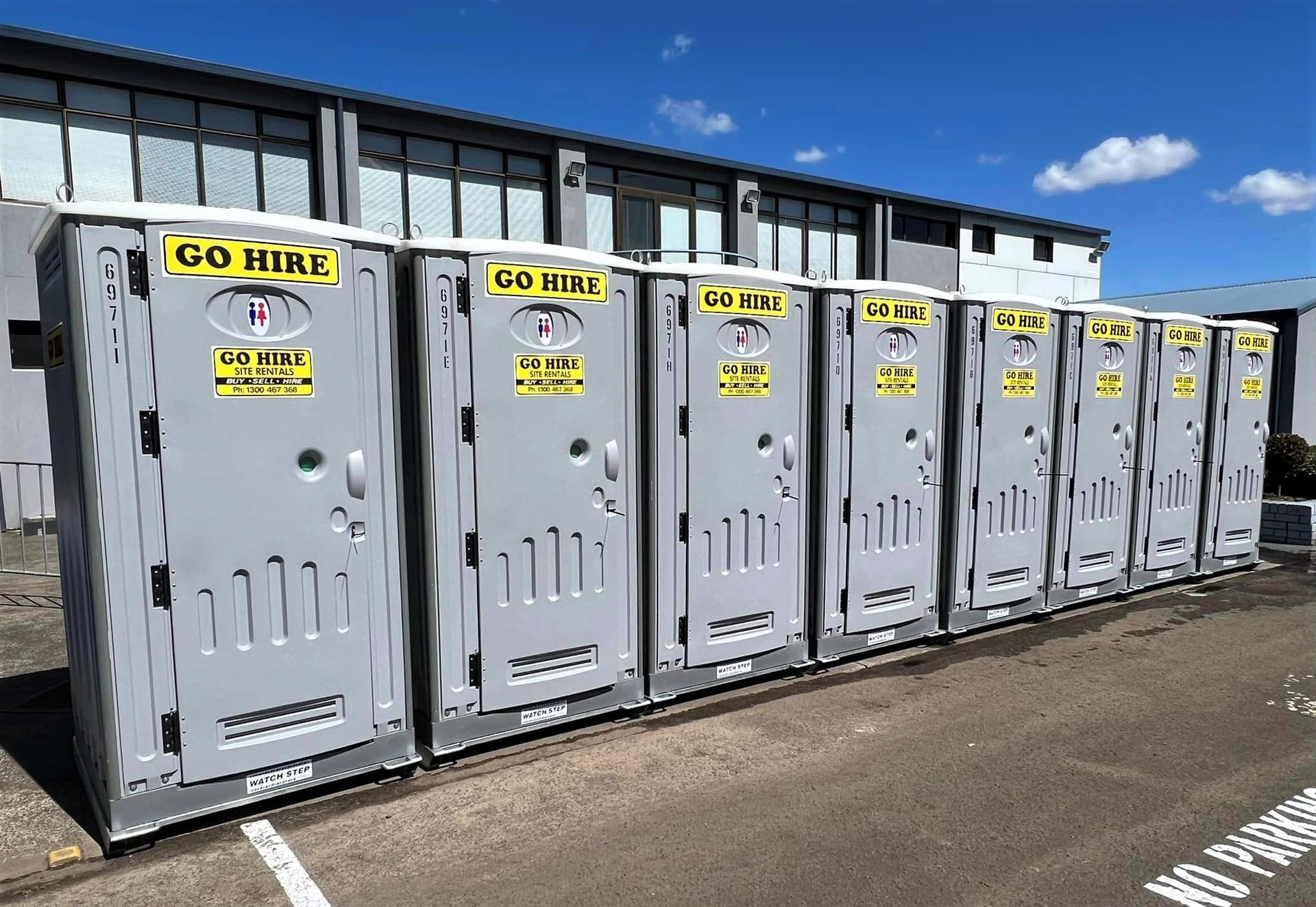 Row of grey portable restroom units lined up outside a commercial building under a clear blue sky. Row of grey portable restroom units lined up outside a commercial building under a clear blue sky.