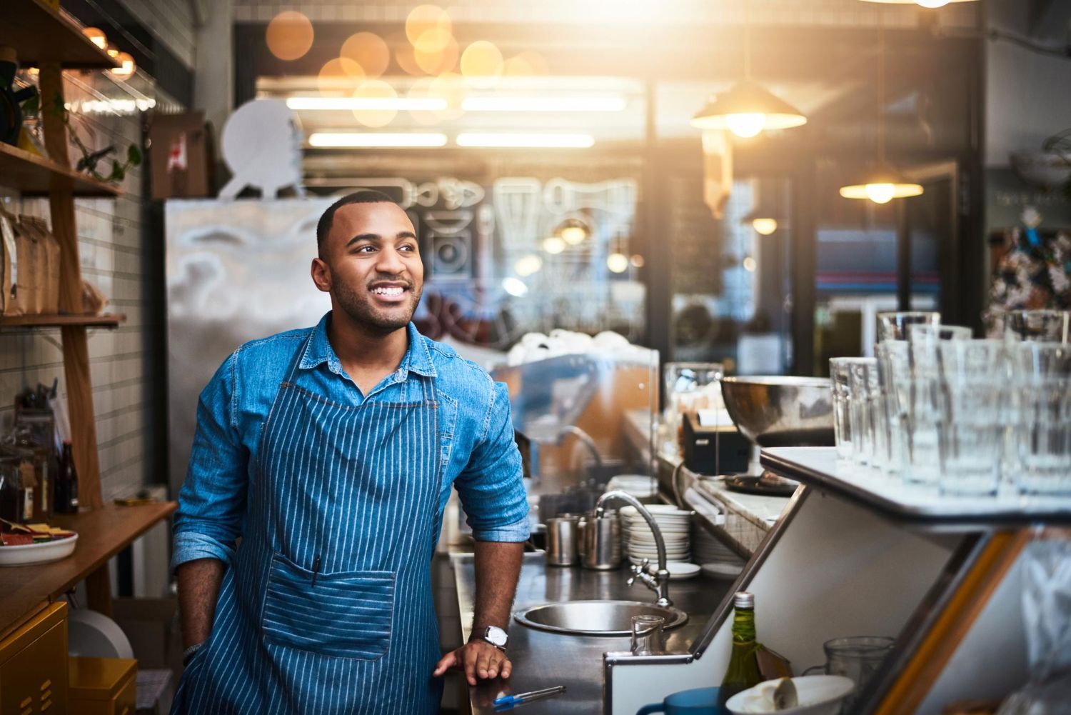 Smiling barista in a blue patterned shirt standing behind a café counter with glassware and coffee equipment.