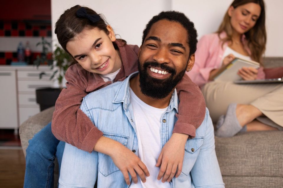 Smiling man with child on his back in a living room, with a woman reading in the background