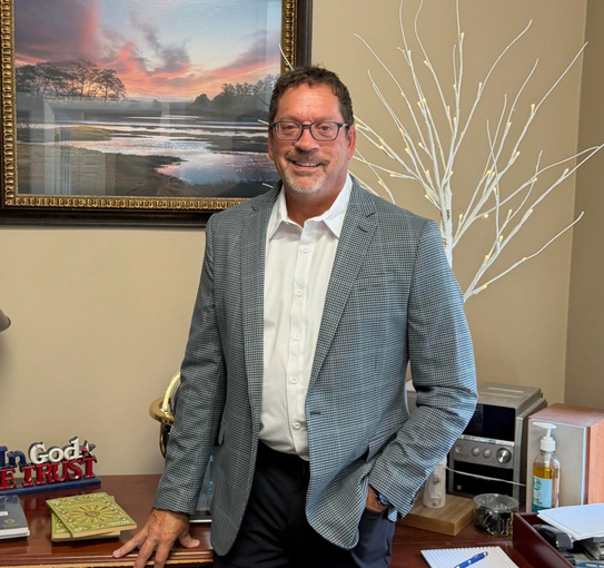 Stephen Podmaska in a checkered blazer standing beside a desk in an office, smiling under framed wall art.