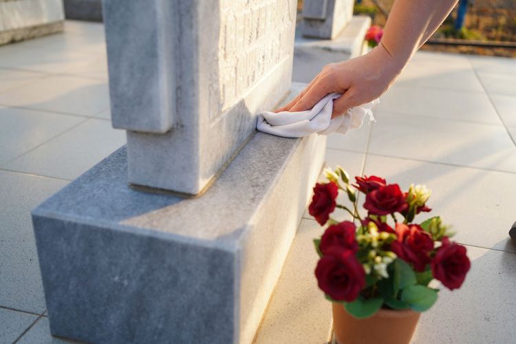 Hand placing a white cloth on a stone memorial beside red flowers in a sunny outdoor setting