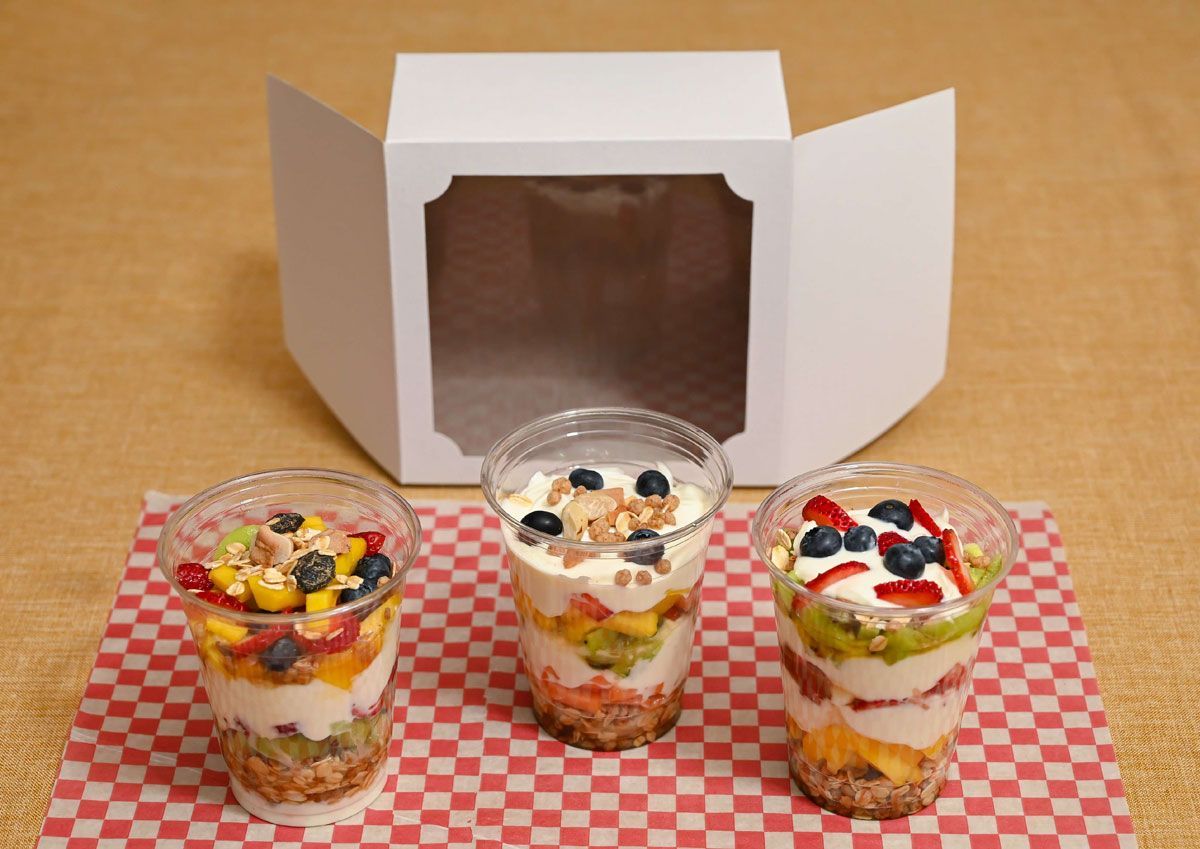 Tres parfaits de frutas y yogur en vasos transparentes sobre un mantel a cuadros rojos, con una caja blanca abierta detrás.