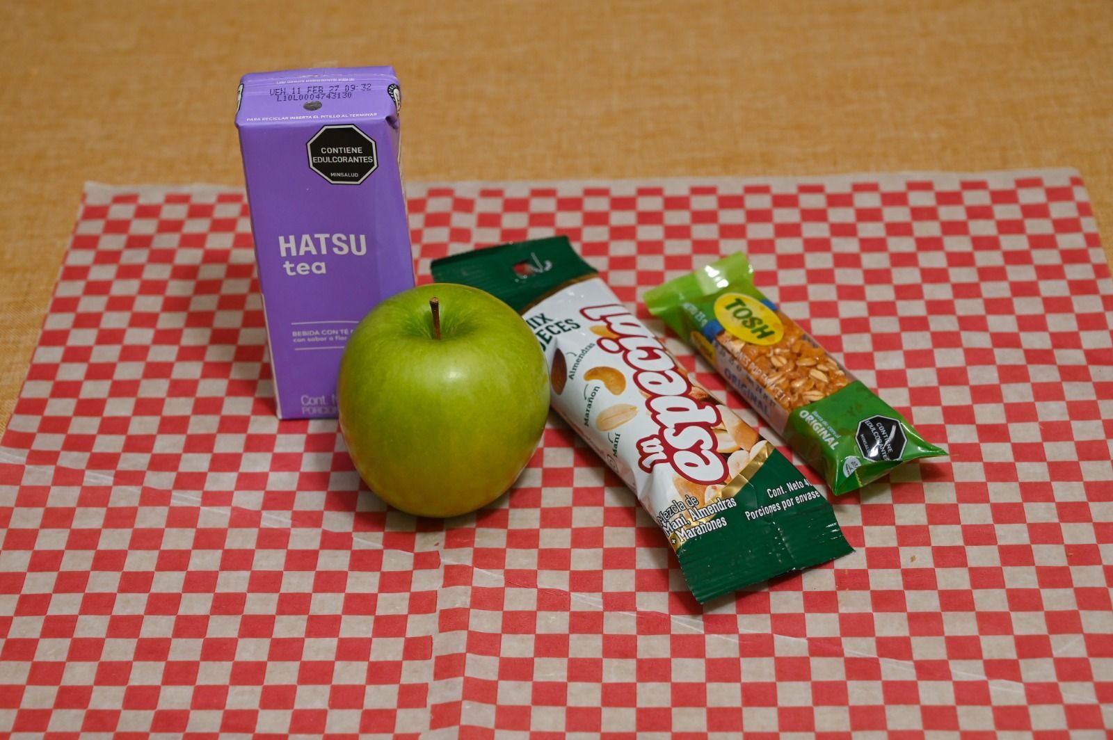Manzanas verdes y aperitivos envasados sobre un mantel a cuadros rojos, con una caja de cartón morada detrás.