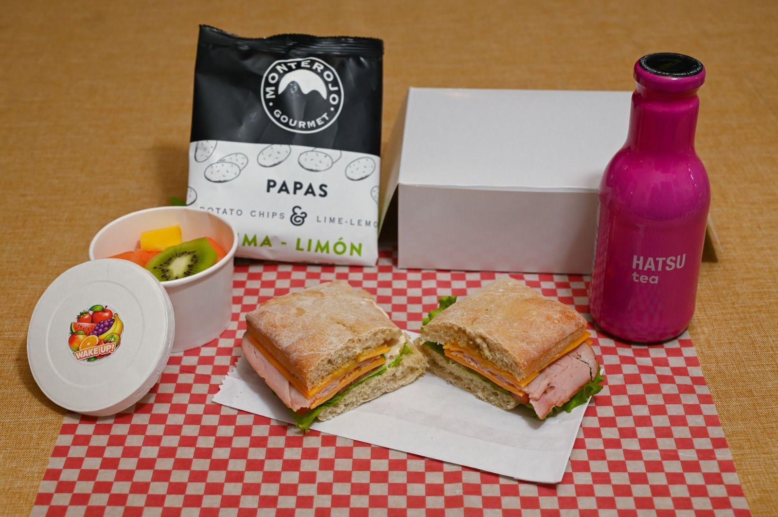 Sándwiches, patatas fritas y una botella rosa sobre un mantel a cuadros con una bolsa de aperitivos en la parte de atrás.