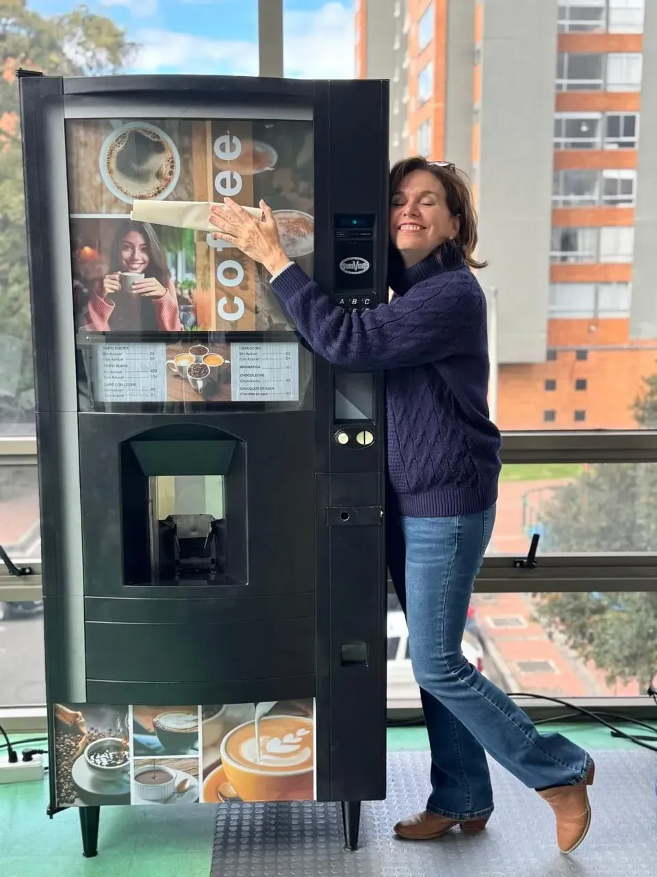 Mujer sonriendo junto a una máquina expendedora negra con imágenes de café, en el interior de una vivienda