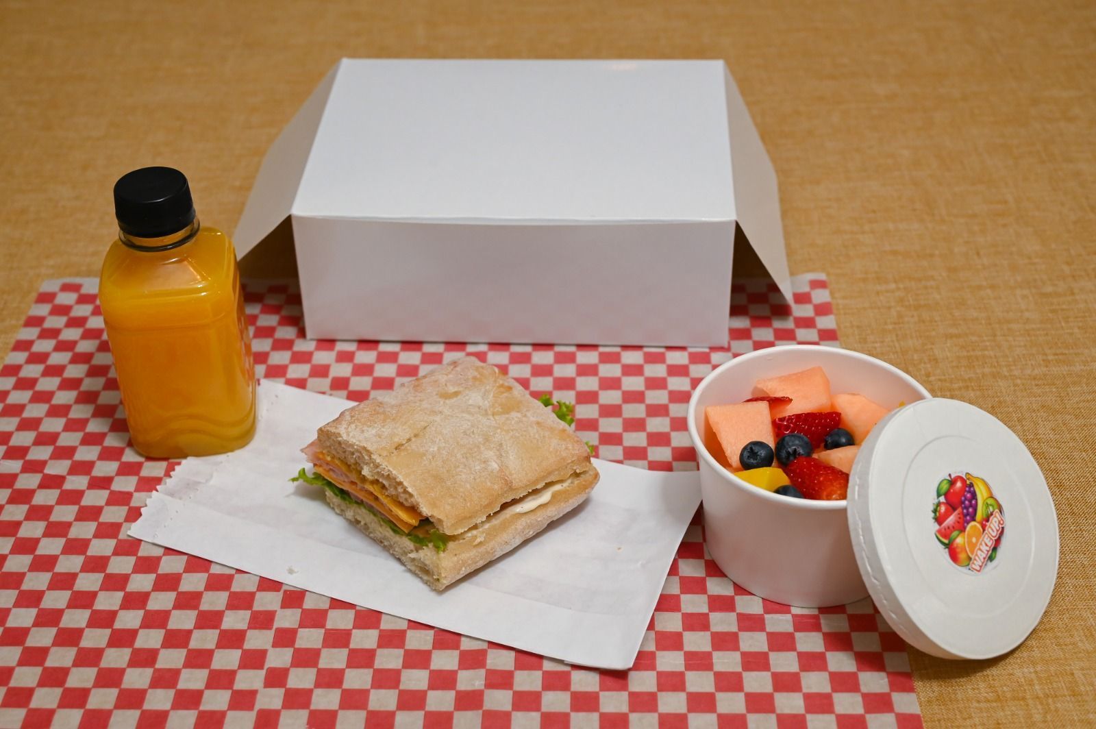 Un sándwich, una botella de jugo y una taza de fruta junto a una lonchera blanca abierta sobre un mantel a cuadros.