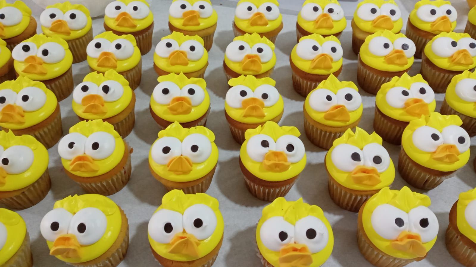 Hileras de cupcakes amarillos con glaseado blanco y ojos de caramelo, dispuestos en una bandeja como pequeñas caritas.