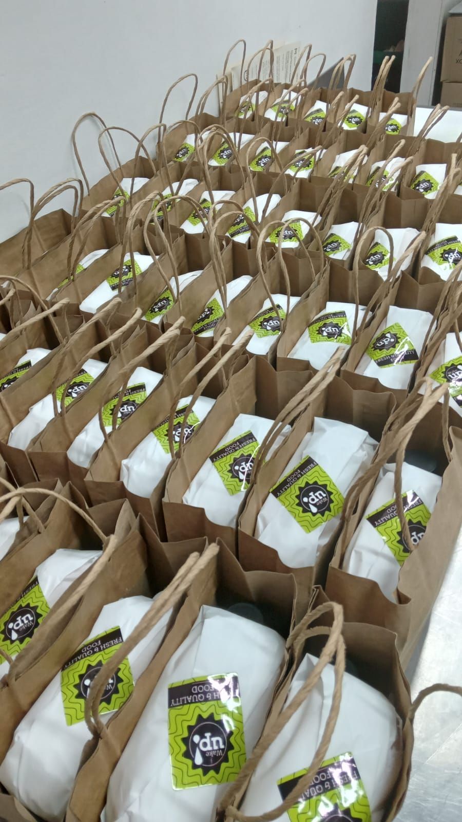 Filas de bolsas de regalo de papel marrón con papel de seda blanco y etiquetas verdes dispuestas sobre una mesa.