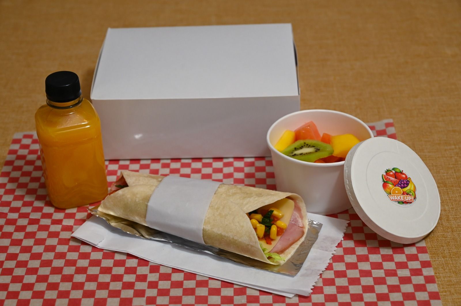 Sándwich empaquetado, bebida de naranja, ensalada de frutas y postre sobre un mantel a cuadros rojos con una caja blanca.