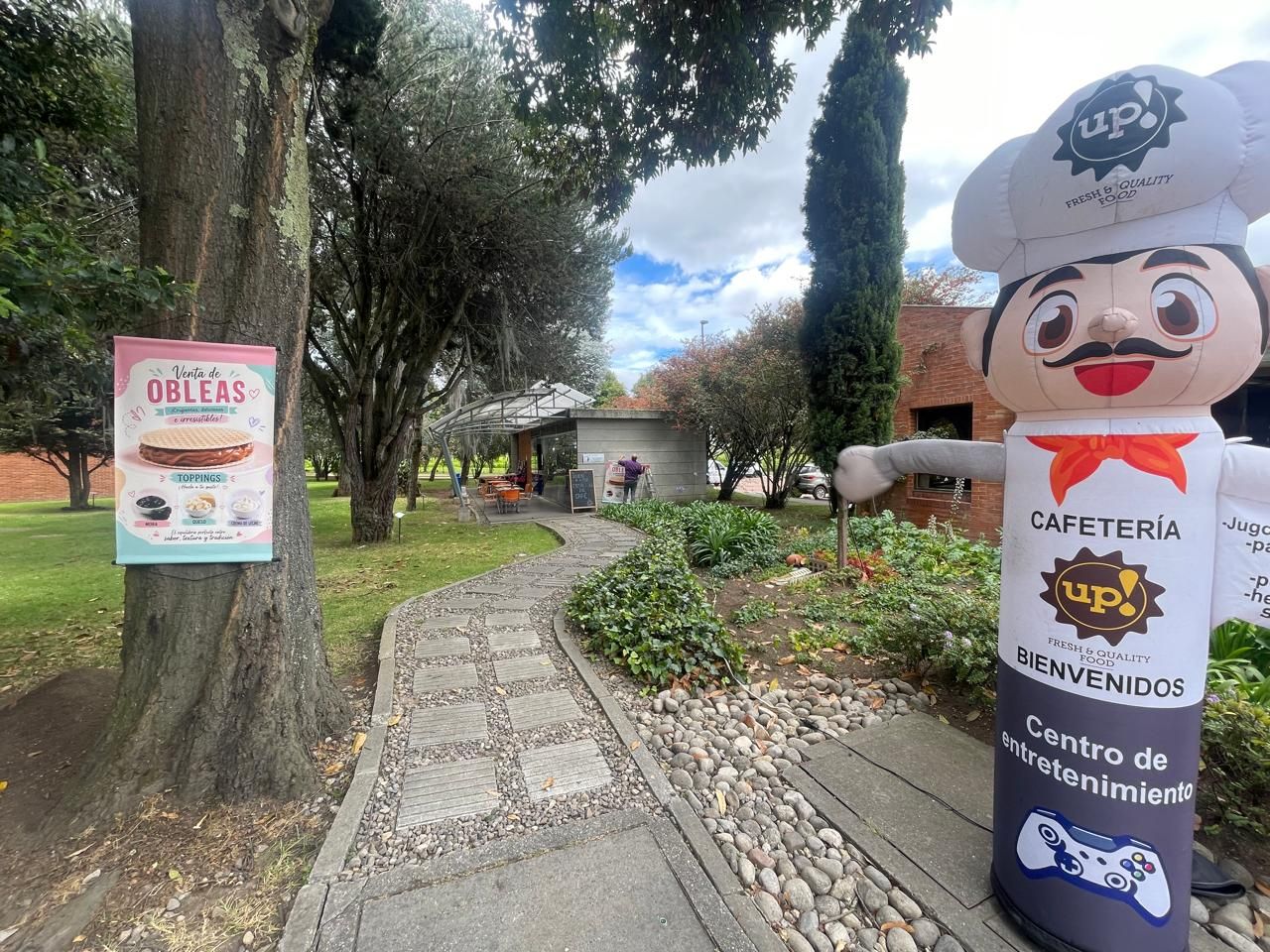 Sendero del parque con pasarela de piedra, árboles y un letrero de restaurante junto a una estatua de un chef