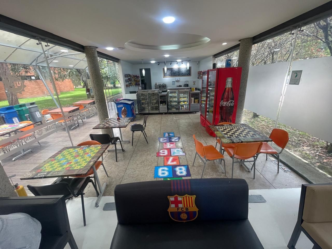 Cafetería exterior cubierta con mesas y sillas coloridas, zona de bar y el escudo del FC Barcelona en el asiento de enfrente.
