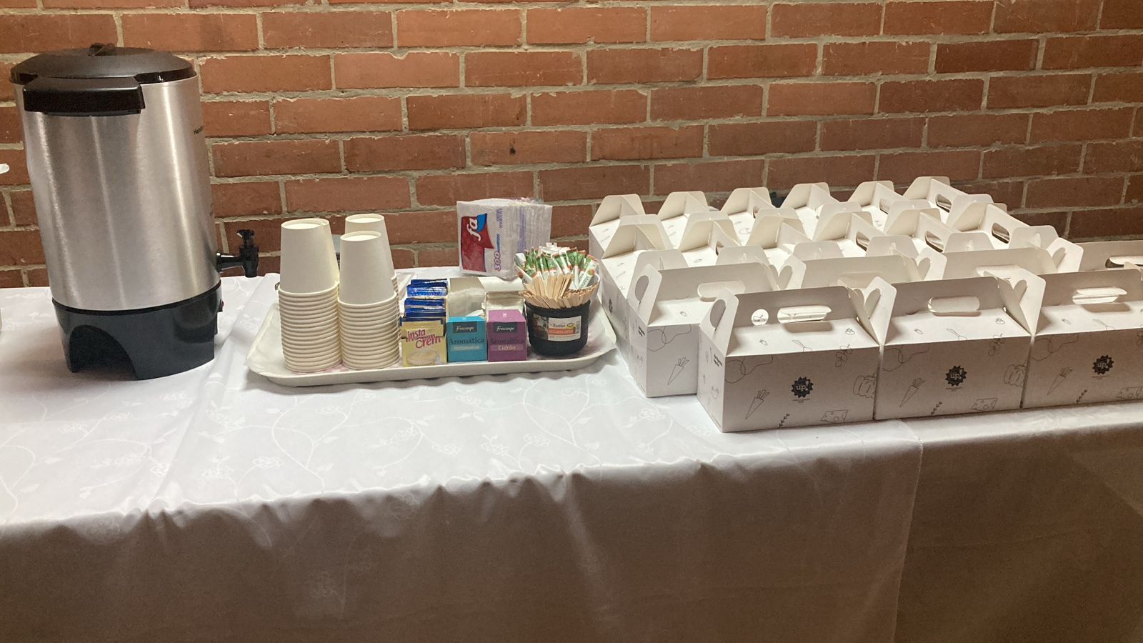 Estación de café y refrigerios con tazas, condimentos y cajas para llevar sobre una mesa contra una pared de ladrillos.