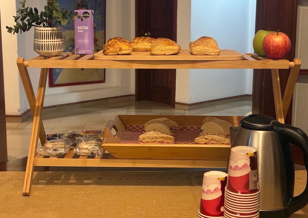 Mesa expositora de panadería con pasteles variados, manzanas, una cafetera y tazas apiladas sobre un suelo alfombrado.