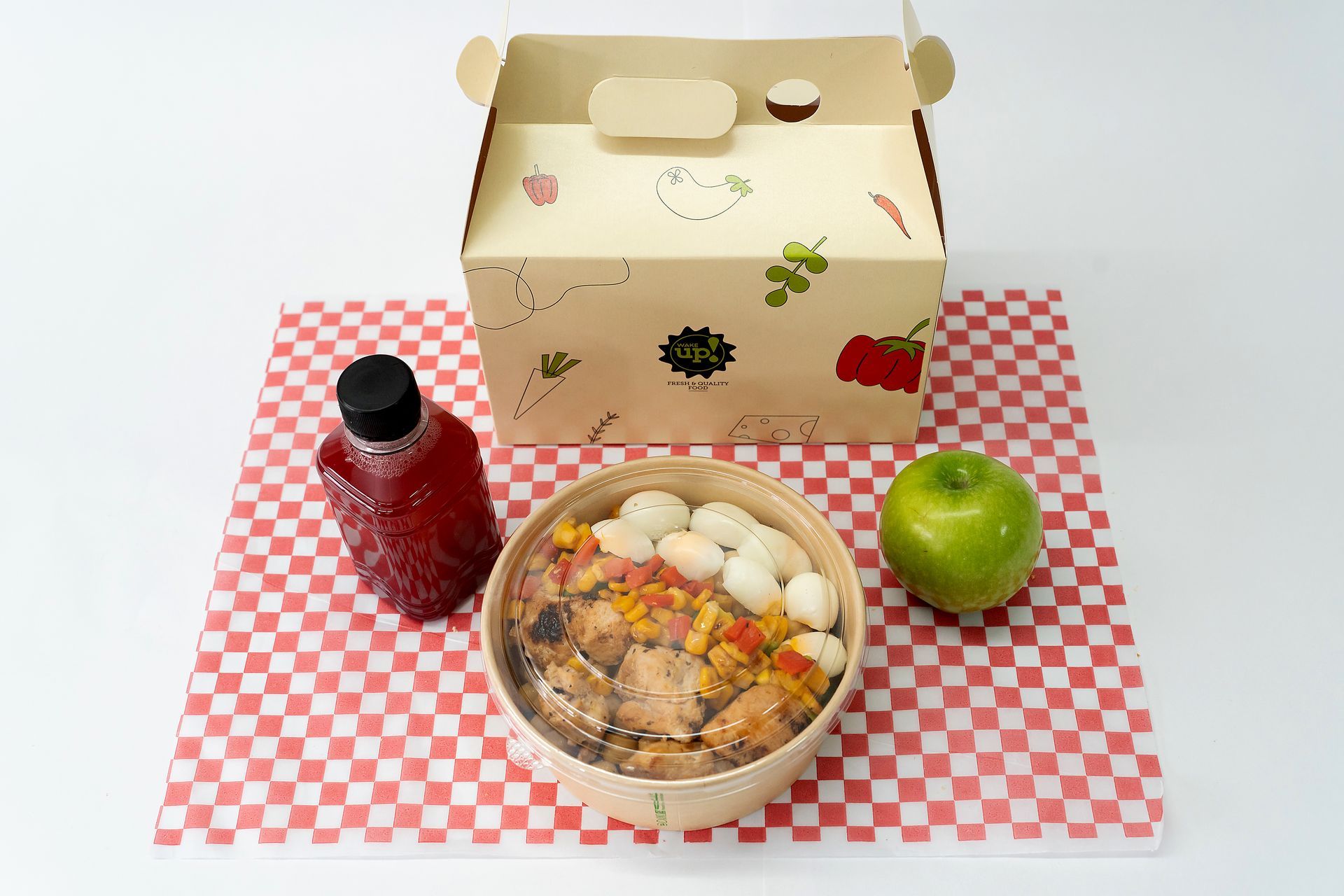 Almuerzo para llevar con ensalada, manzana, botella de bebida roja y una lonchera decorada sobre un tapete a cuadros.