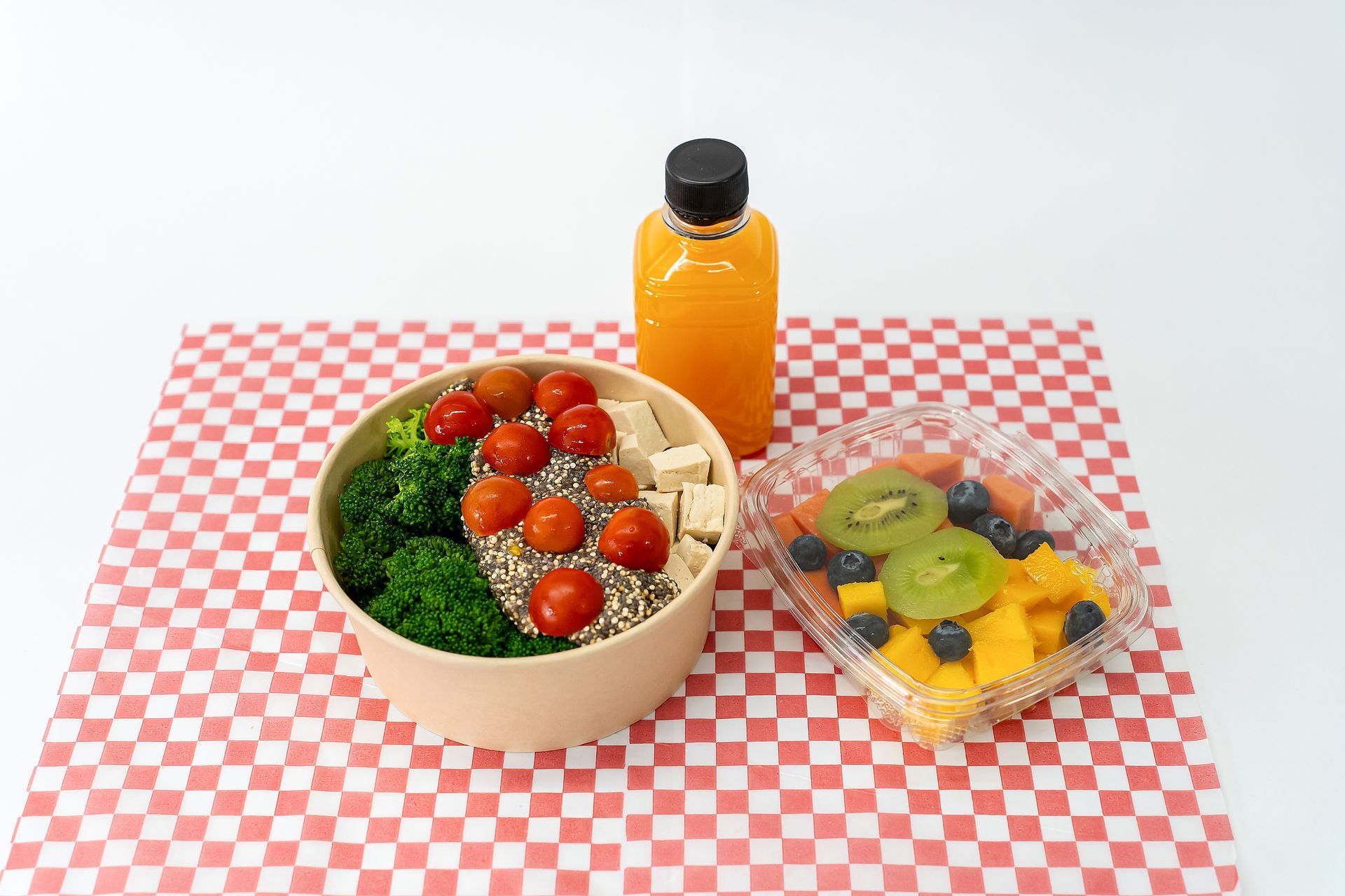 Ensaladera, bebida embotellada y vaso de fruta sobre un mantel a cuadros rojos y blancos.