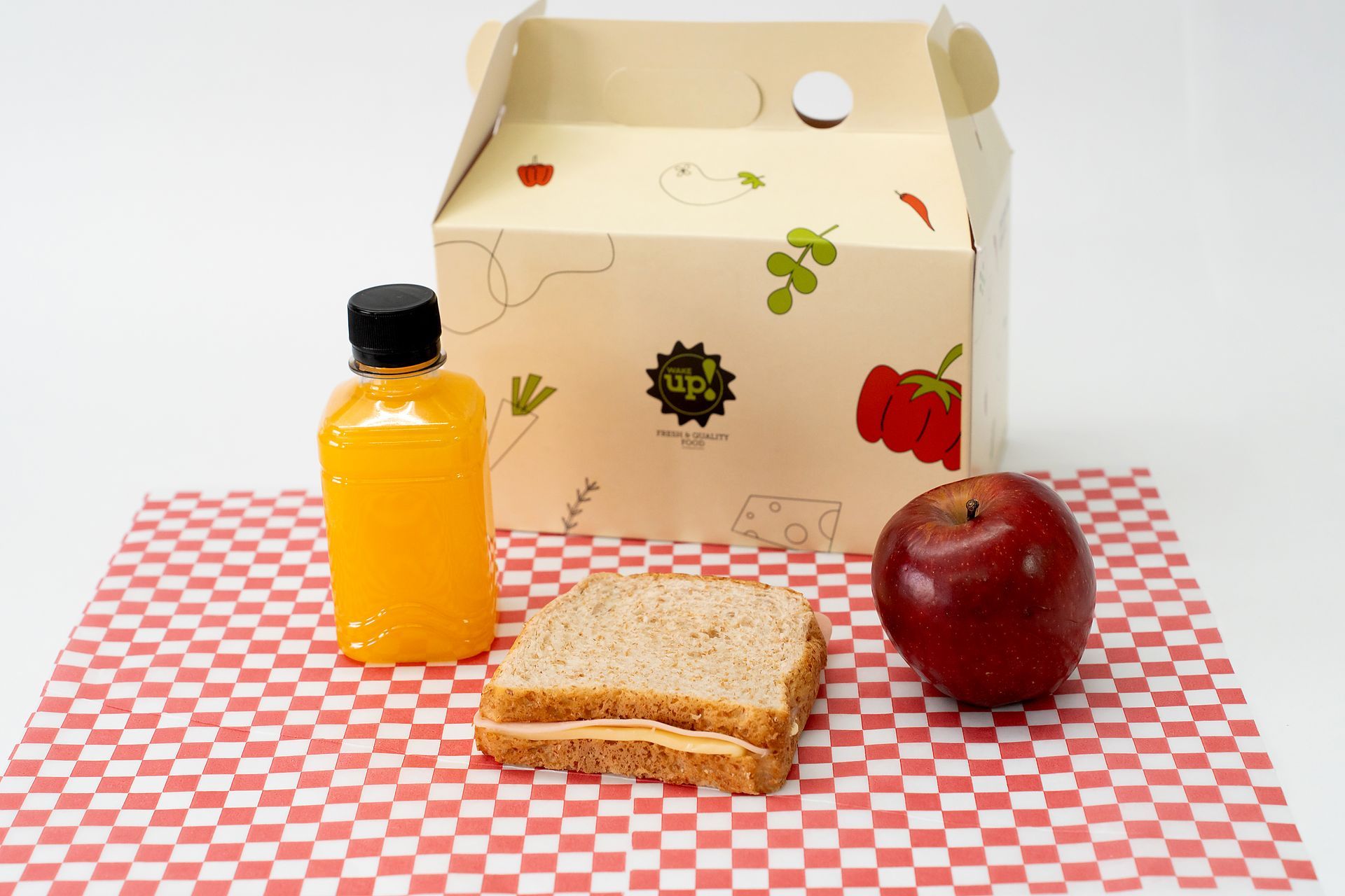 Artículos para picar sobre una mesa a cuadros rojos: zumo de naranja, sándwich, manzana y un fondo de cartón.