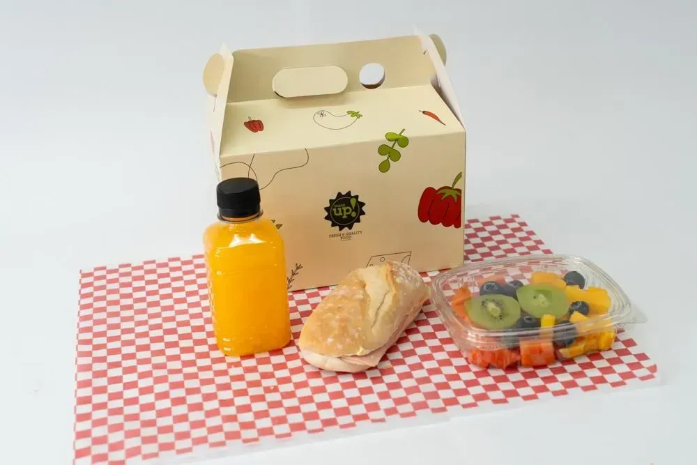Artículos para la lonchera sobre un paño a cuadros rojos y blancos: botella de jugo, sándwich y ensalada de frutas.