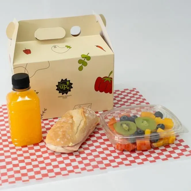 Fiambrera preparada con bebida de naranja, panecillo y tarrina de fruta sobre un mantel a cuadros.