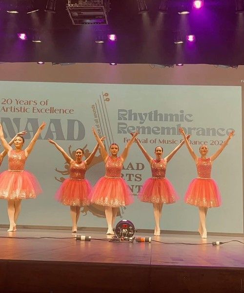 Five dancers in pink tutus on stage with arms raised under purple lights during a recital.