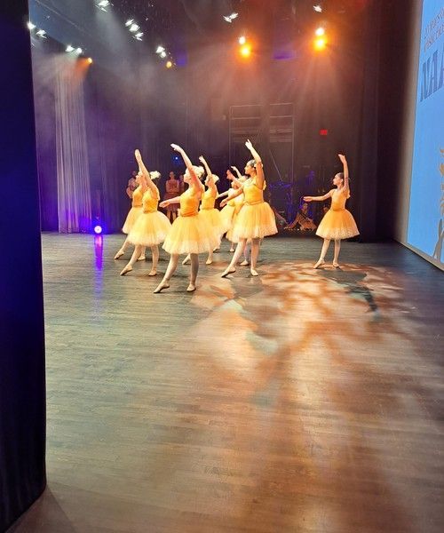 Dancers in yellow dresses perform on a theater stage under warm spotlights.
