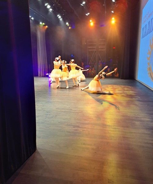Ballet dancers in yellow dresses perform on a theater stage with warm spotlights.
