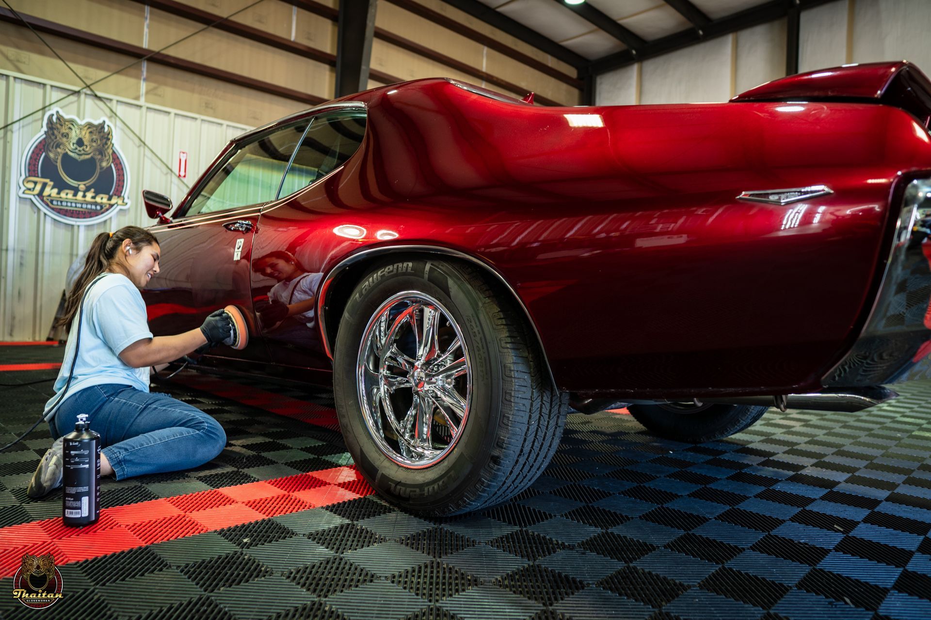 Woman polishing a classic red car in a garage with black and red tiled floor.