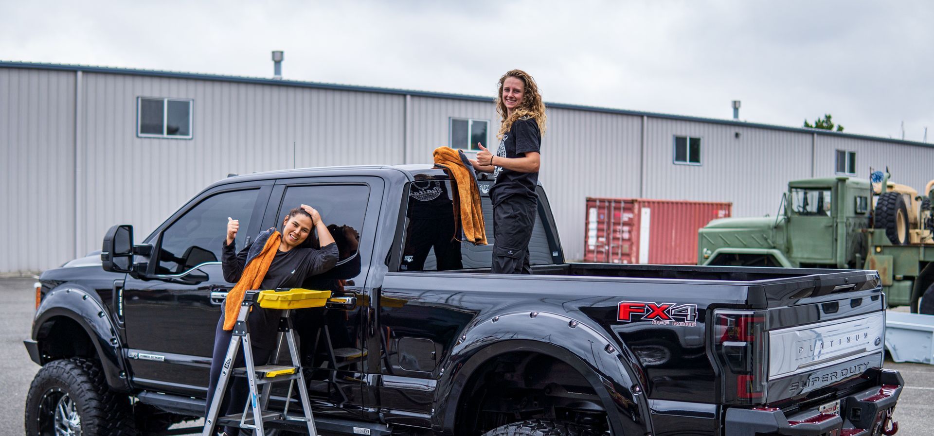 Two people on a black truck. One in the bed, one on a ladder. Industrial setting with military vehicles.
