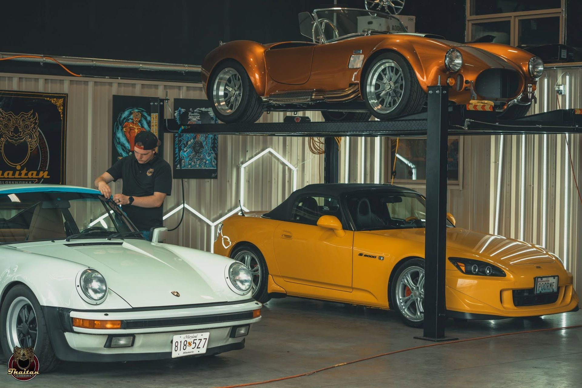 Three sports cars in a garage, with an orange car lifted above a white and a yellow car.