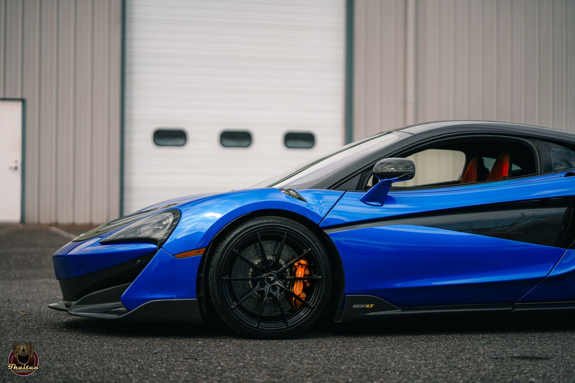 Blue McLaren sports car parked in front of a building, black wheels, orange brake calipers.