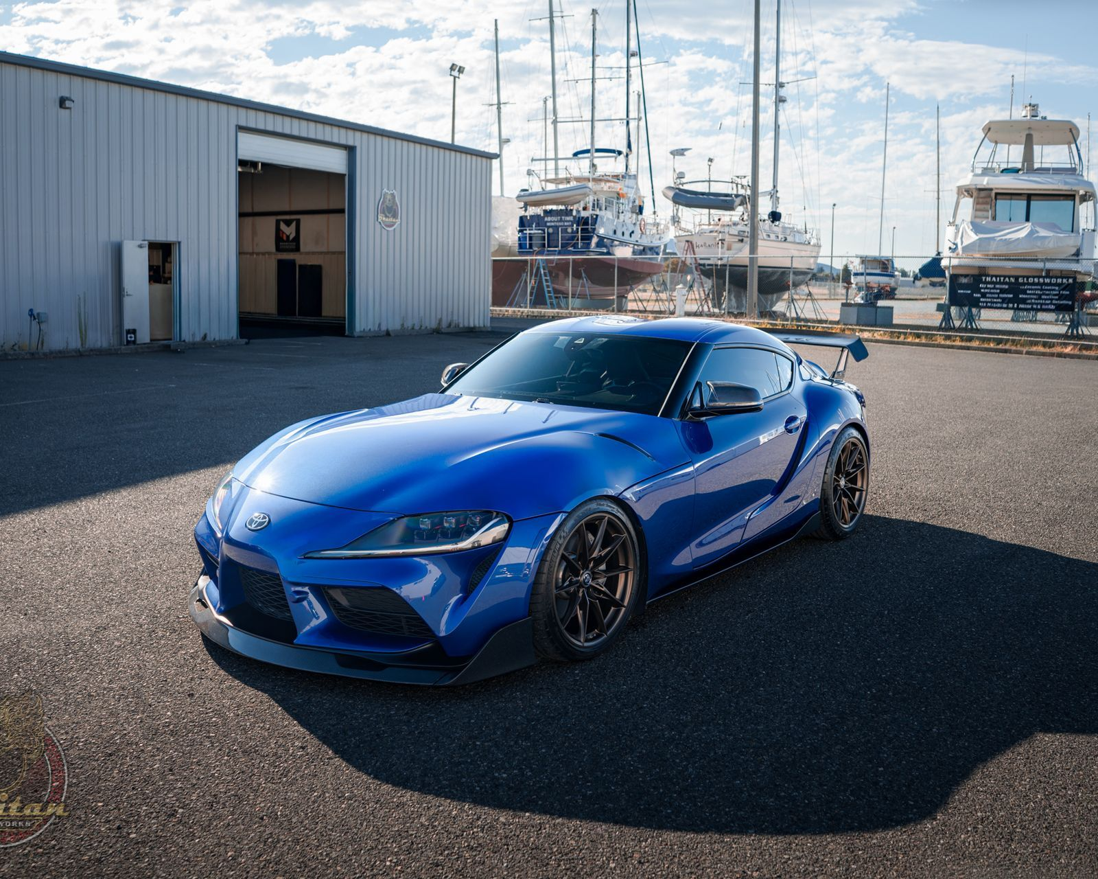 Blue sports car parked on a dock near a marina, with boats and a warehouse in the background