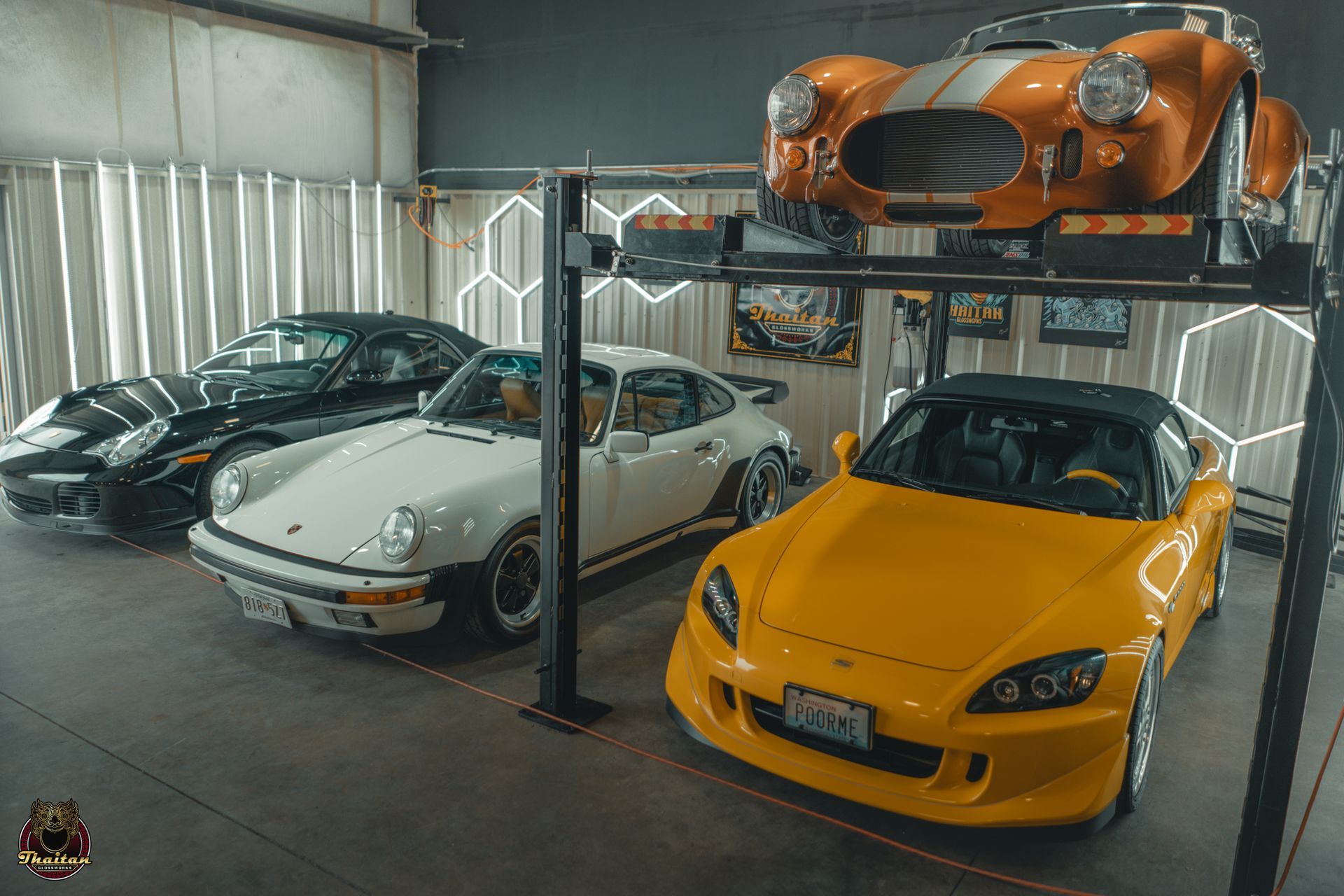 Garage with classic cars: orange Cobra, black Corvette, white Porsche, yellow Honda, & car lift.