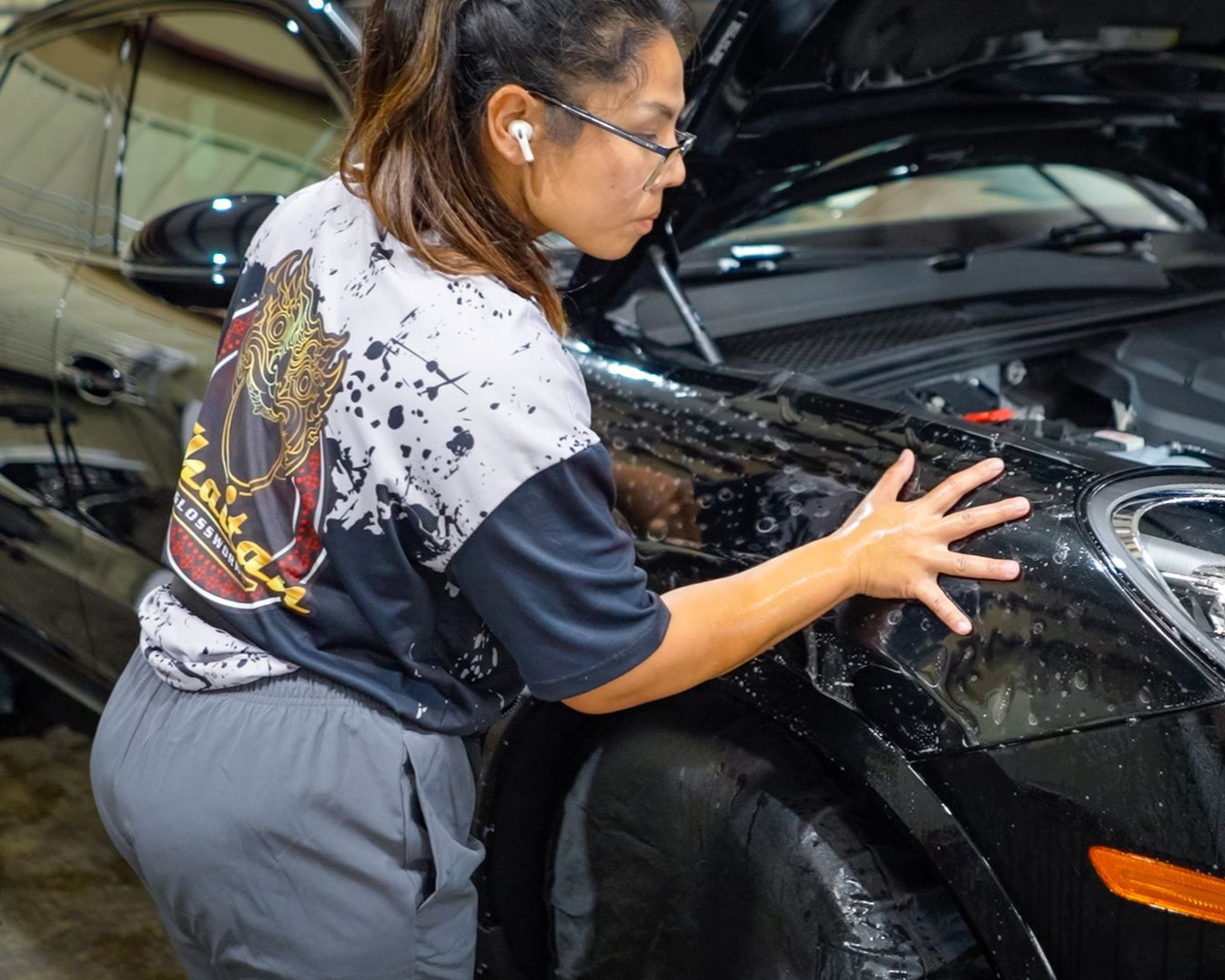 Person polishing the front fender of a black car in a garage, with the hood open.