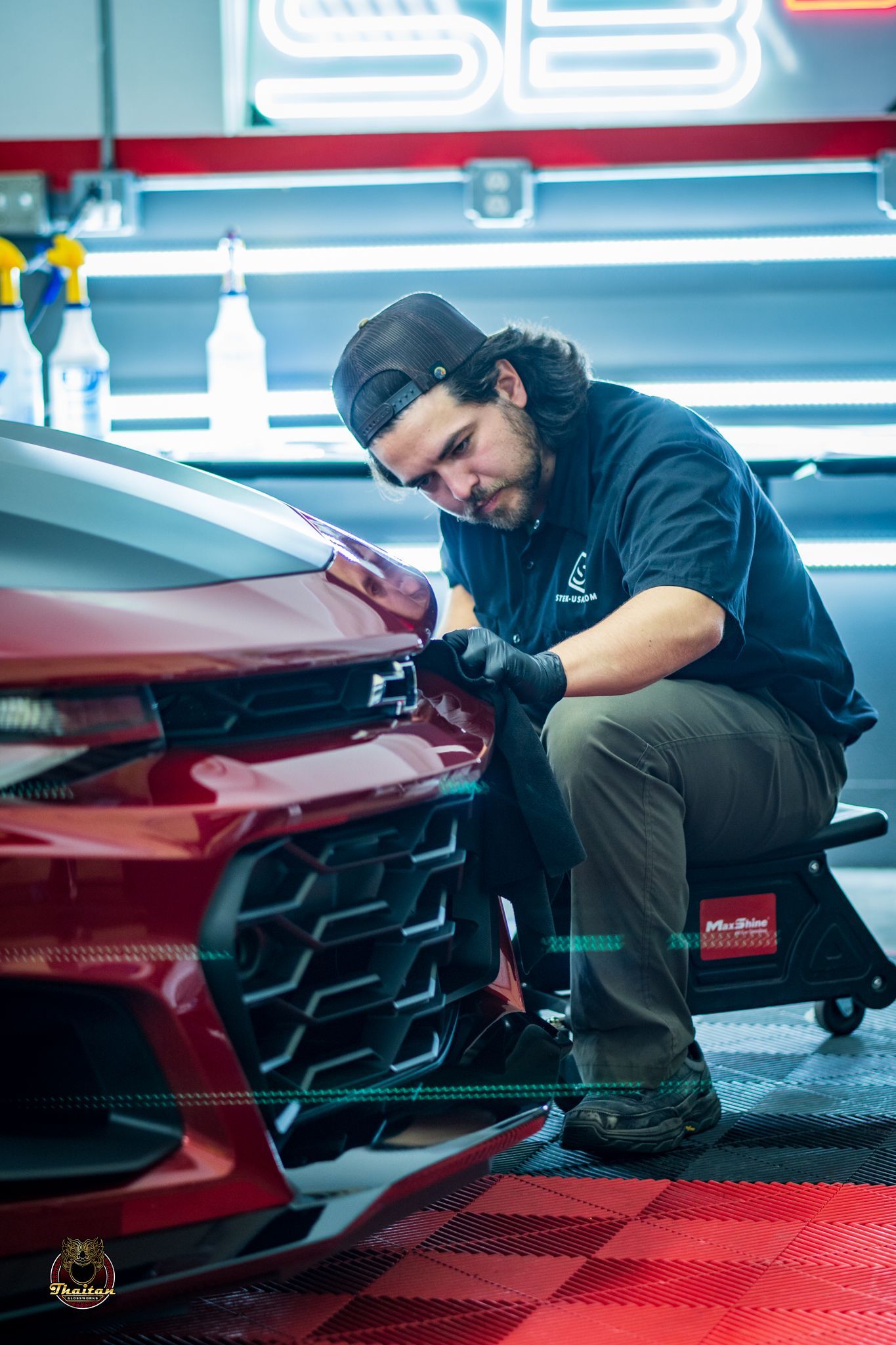 Man in black shirt working on a red car's front grill with tools in a shop.