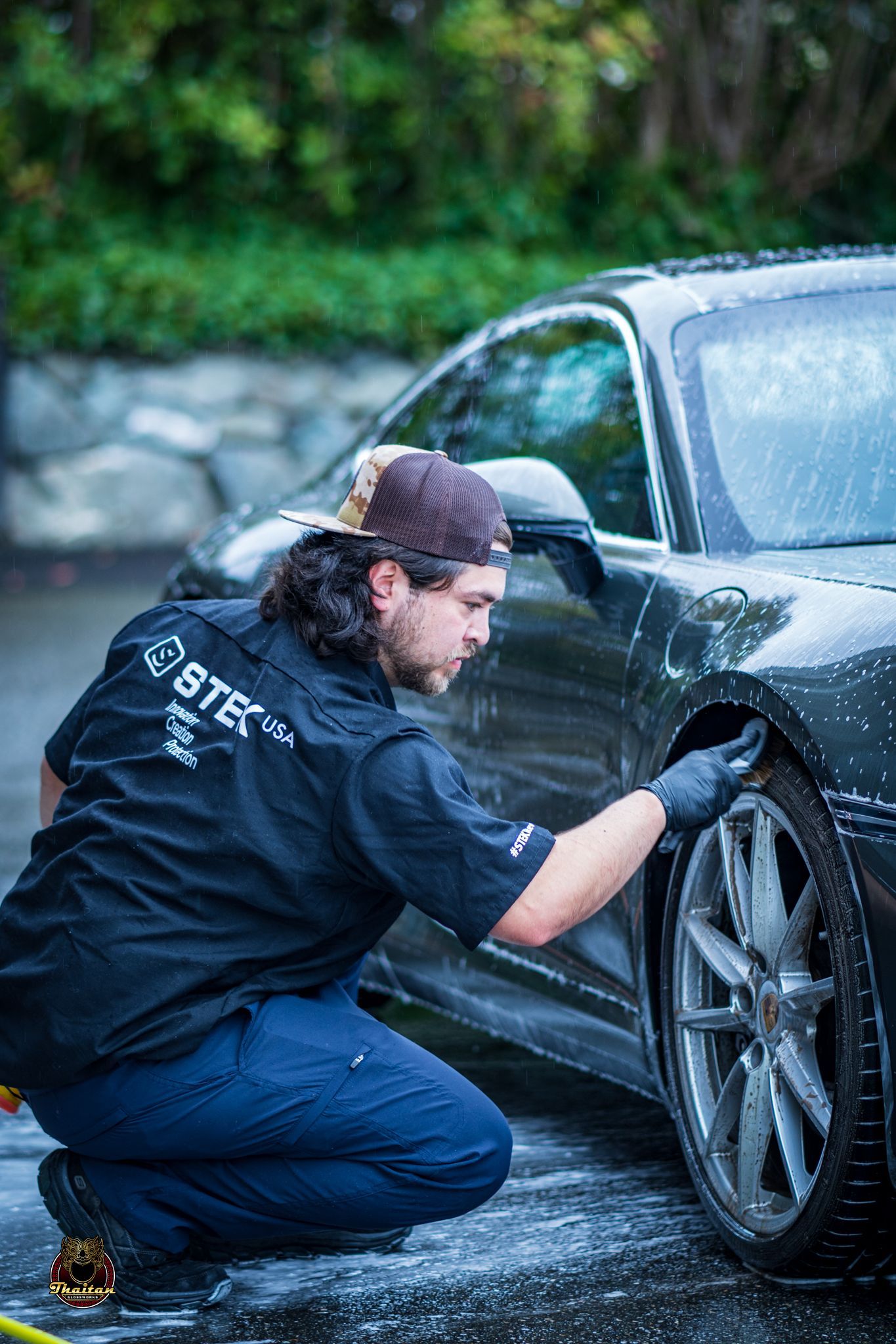 Man washing car tire with black gloves, dark clothing, and a cap outdoors.