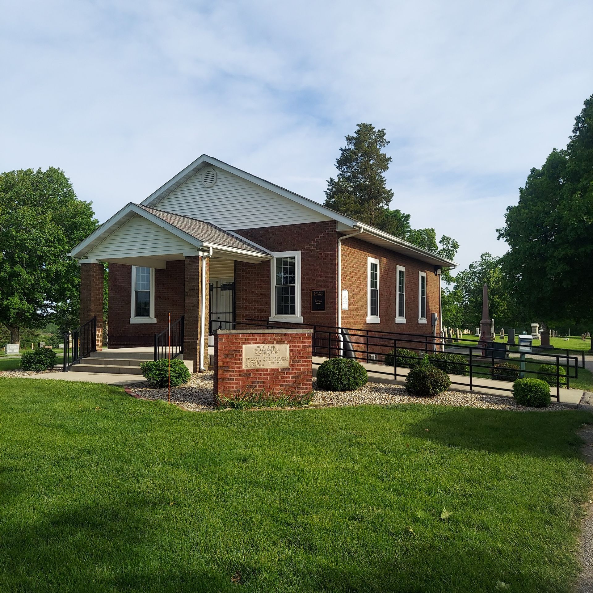 Small brick building with white trim and a covered porch on a grassy lawn under a partly cloudy sky