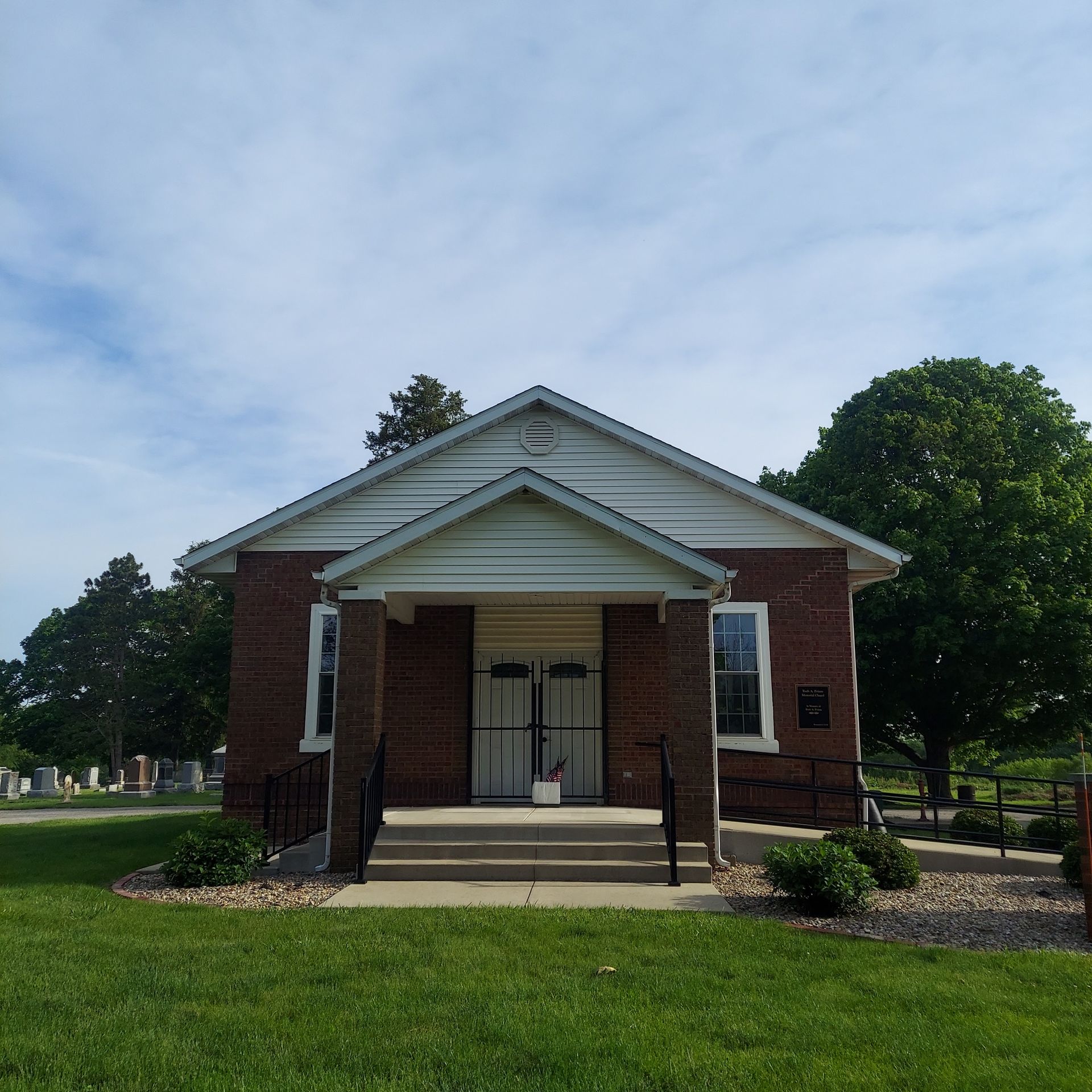 Small brick building with white trim and a front porch, set in a grassy park under a blue sky
