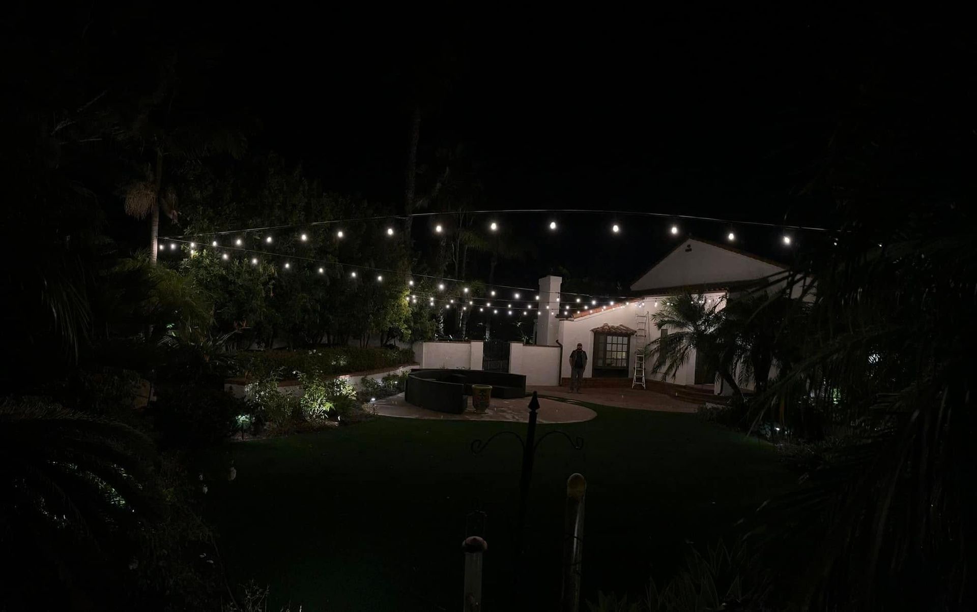 Dim garden patio at night with string lights, white lounge chairs, and a lit umbrella under trees
