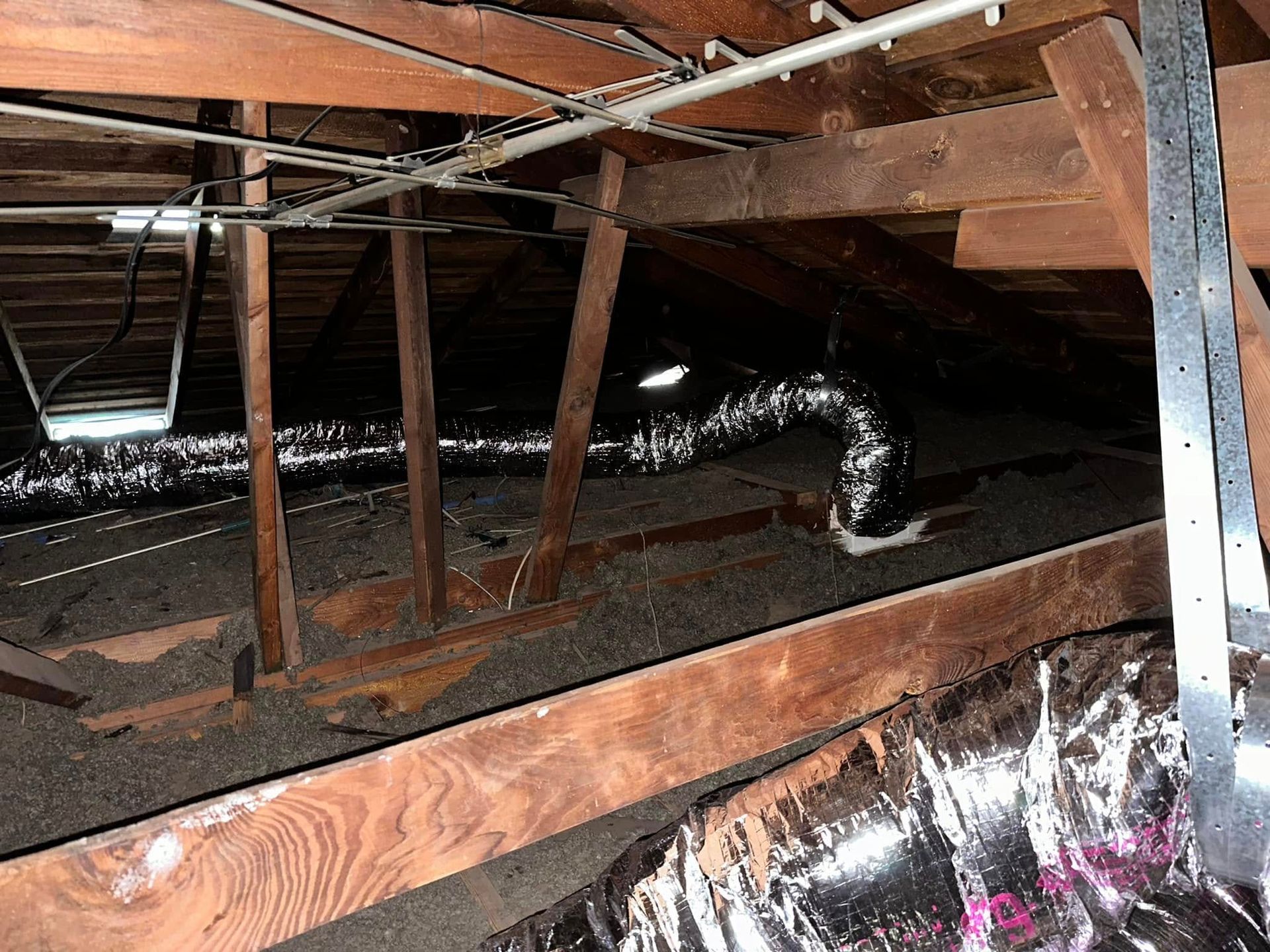 Exposed attic with wooden beams, metal ducting, and insulation in dim light