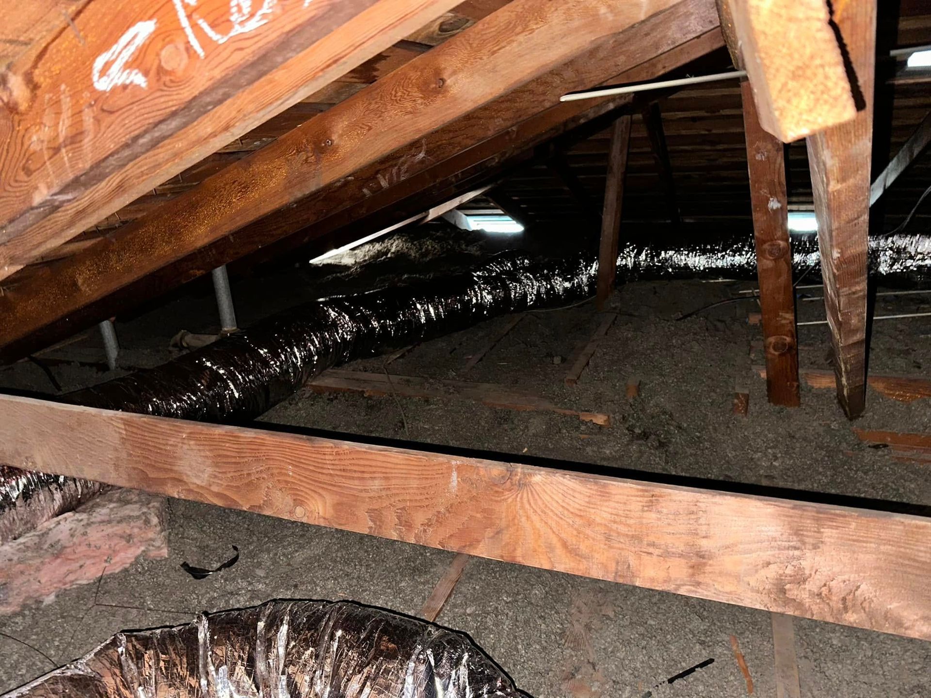 A wooden crawlspace with exposed beams, dark plastic liner, and dirt floor under a slanted structure