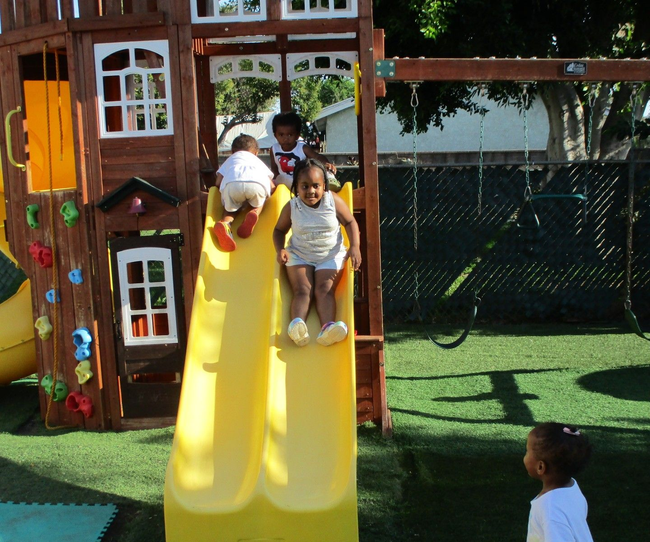 Children sliding down a bright yellow slide at a playground beside a wooden playhouse