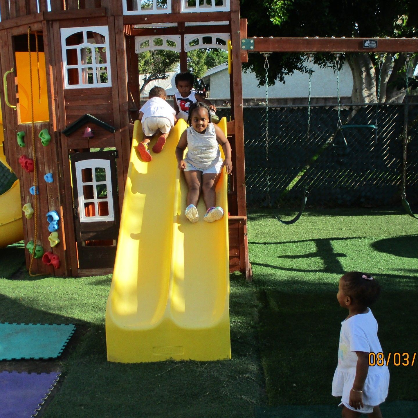 Children on a yellow playground slide in a sunny play area, with another child standing nearby.