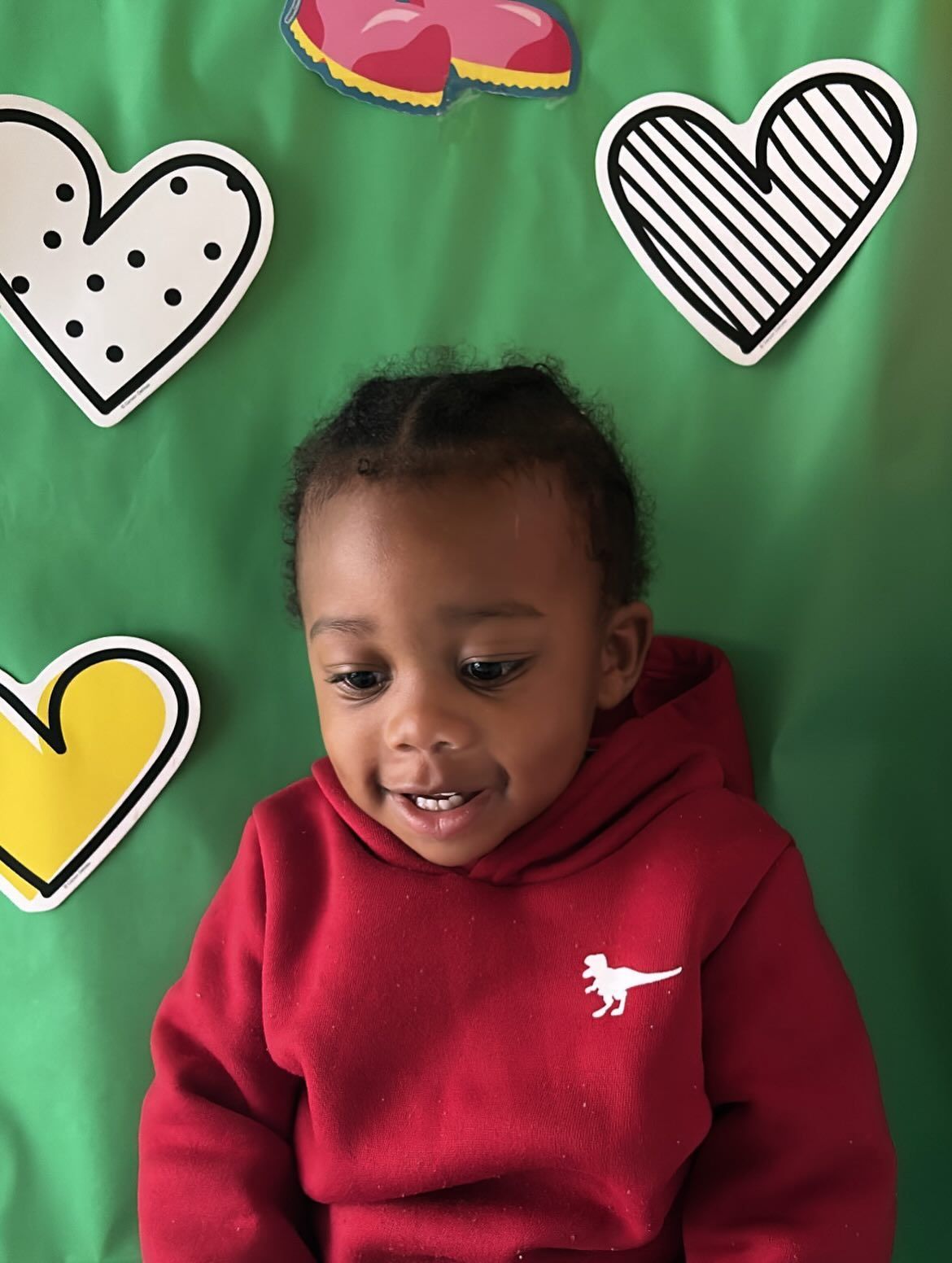 Child in a red hoodie in front of a green background with heart decorations, looking down and smiling