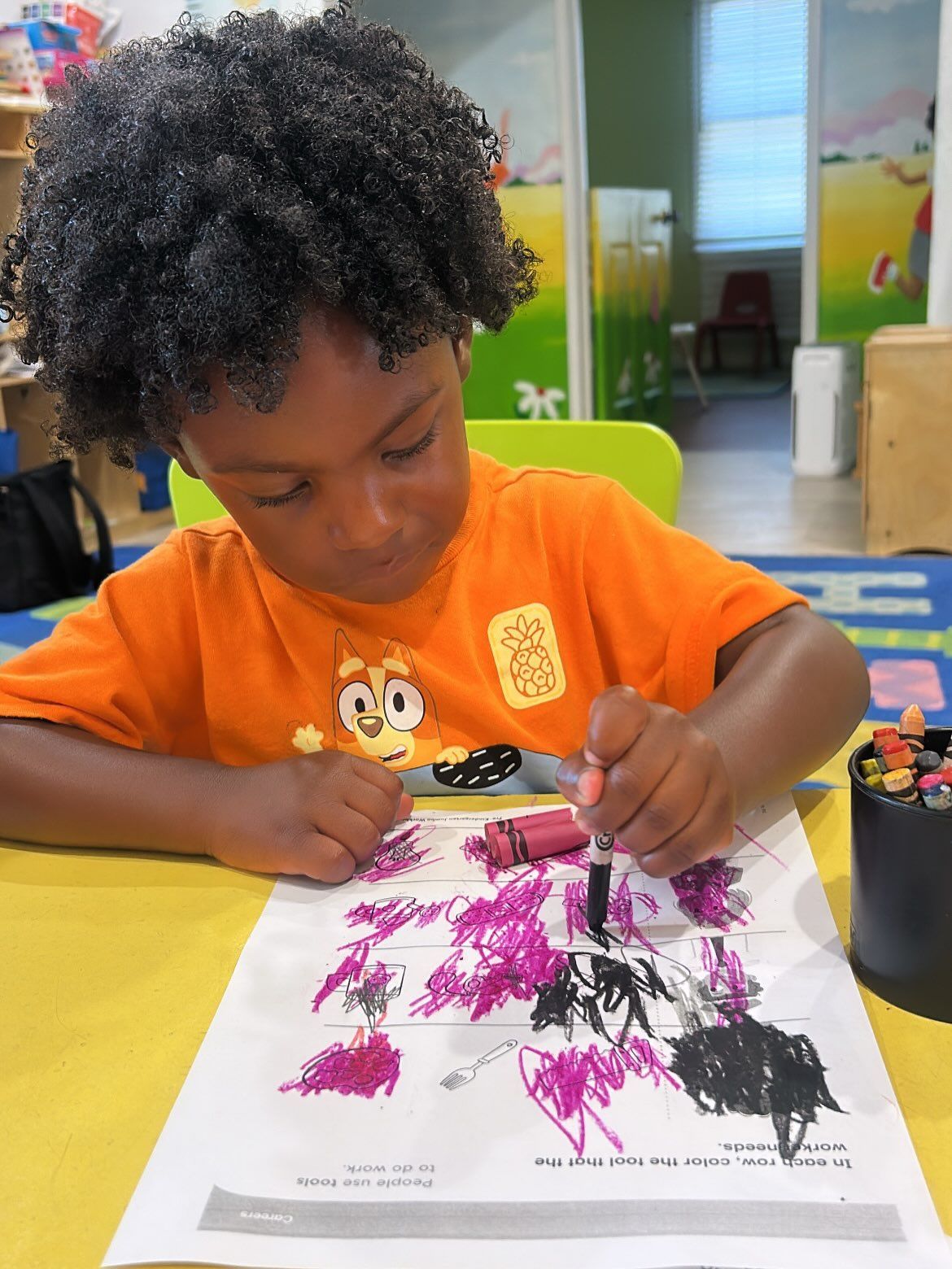 Child in an orange shirt paints with a brush on paper at a table in a classroom.
