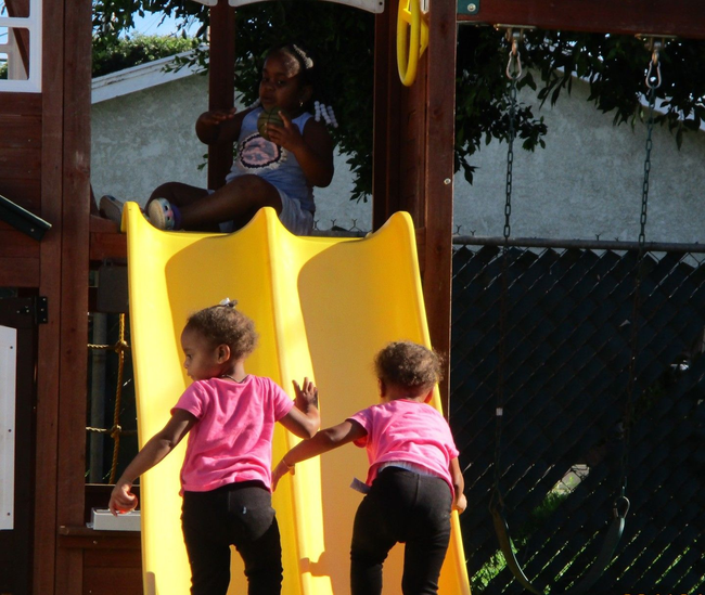 Two children in pink shirts climbing a yellow playground slide while an adult sits above them