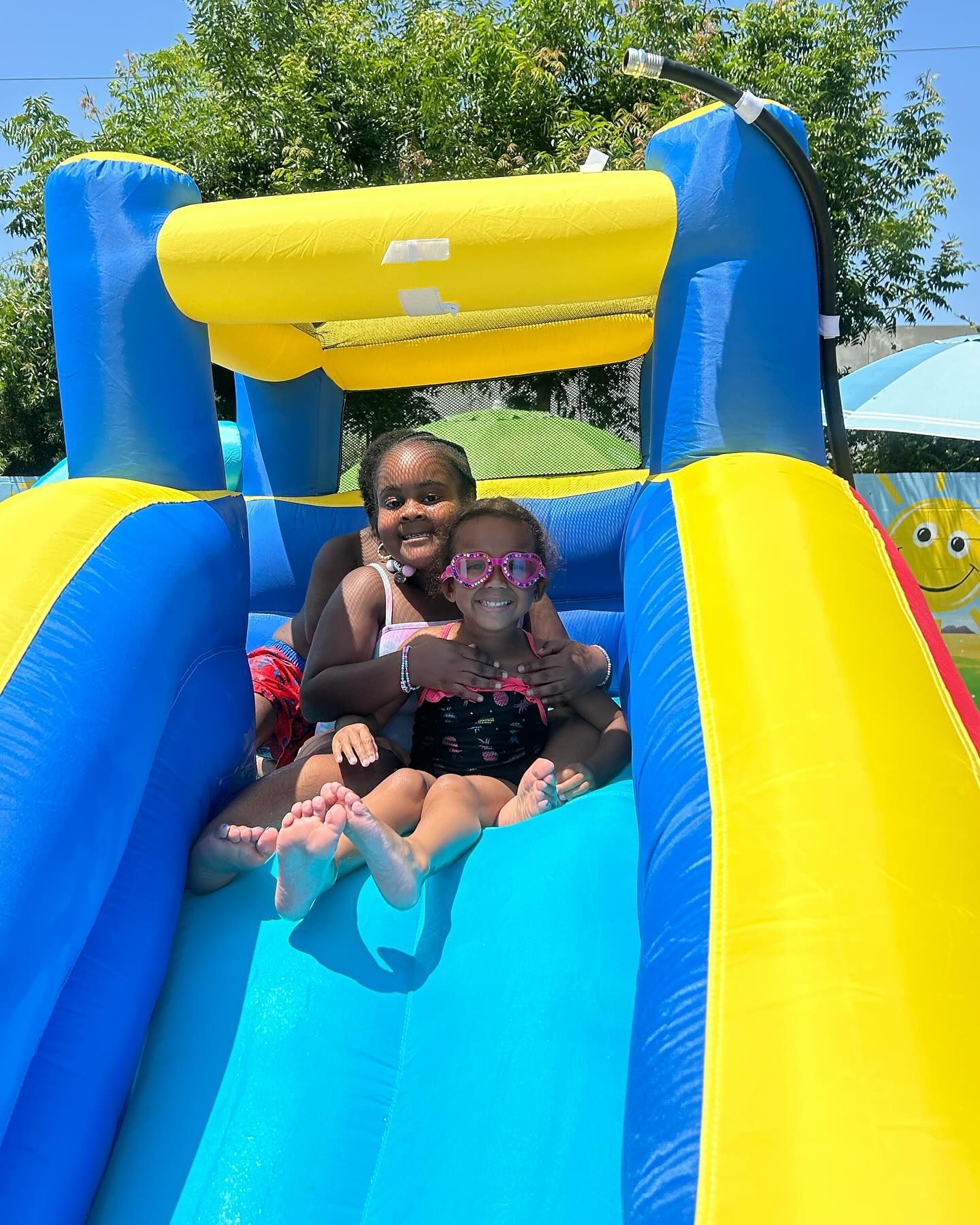 Two children smiling on a blue and yellow inflatable slide on a sunny day.