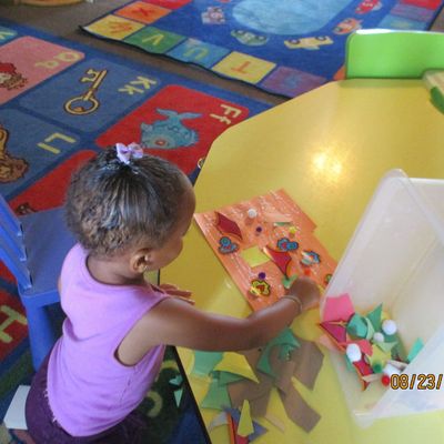 Child arranging colorful shapes on a table in a bright classroom with patterned rug and slide nearby
