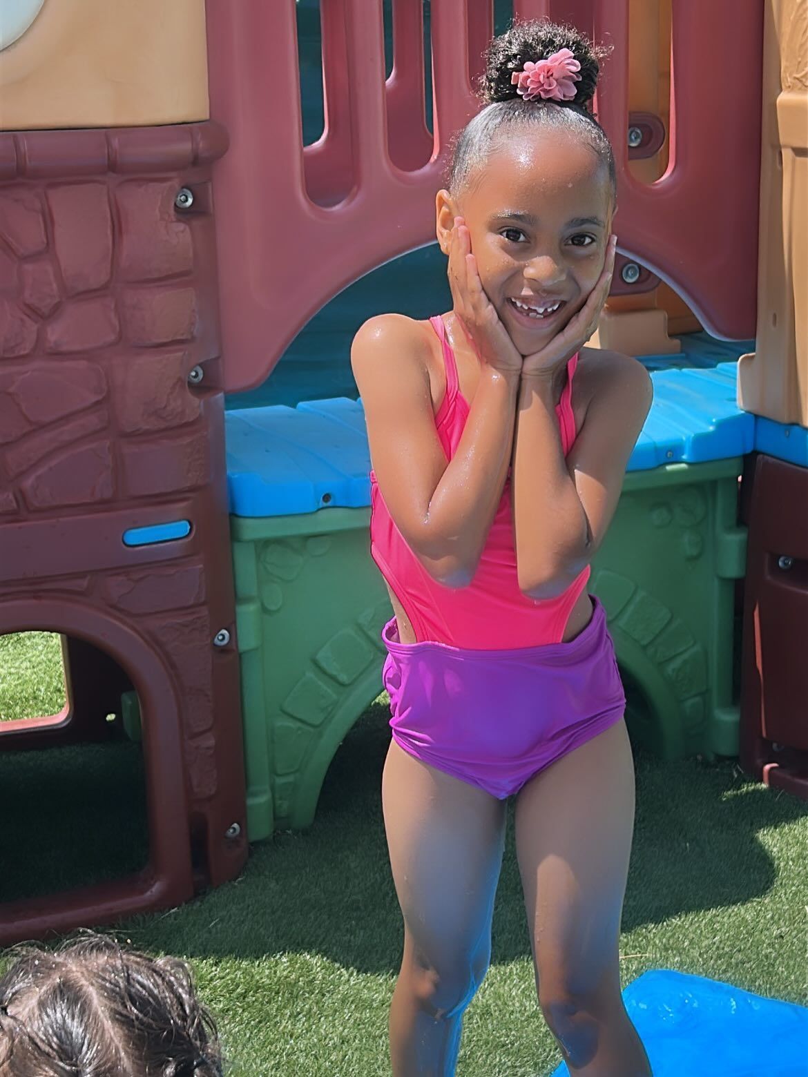 Smiling child in a pink swimsuit posing by a colorful playground structure.