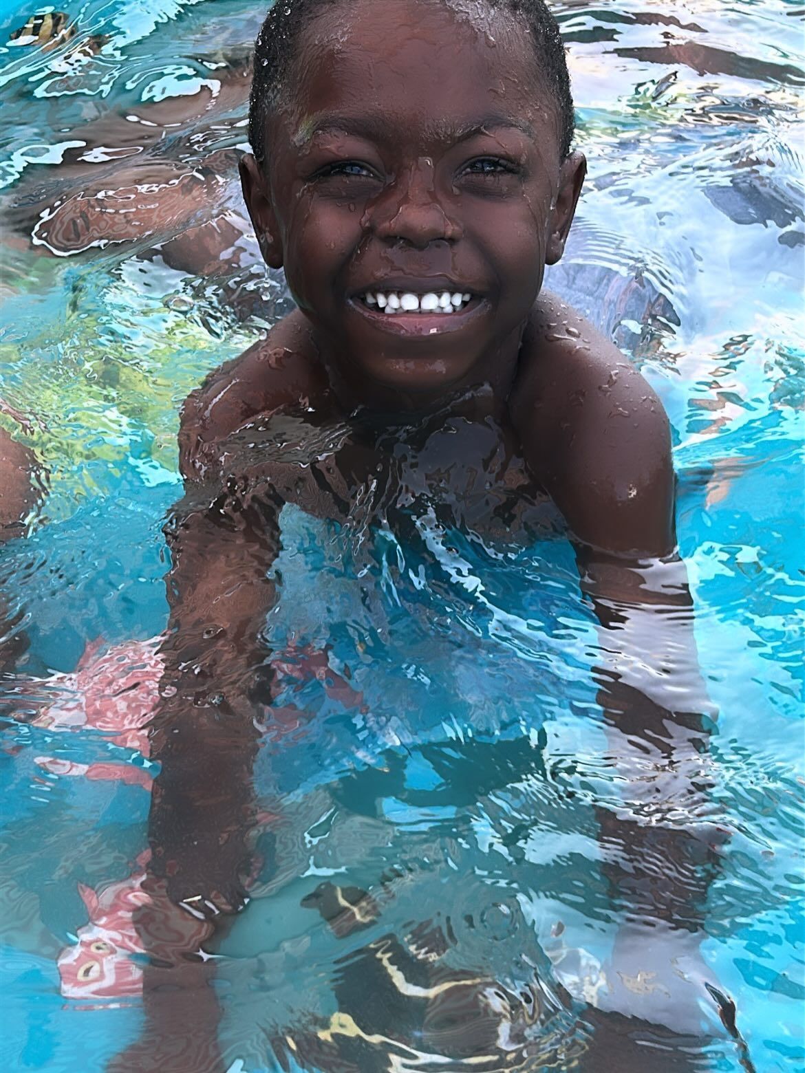 Child smiling in a swimming pool, splashing water toward the camera.