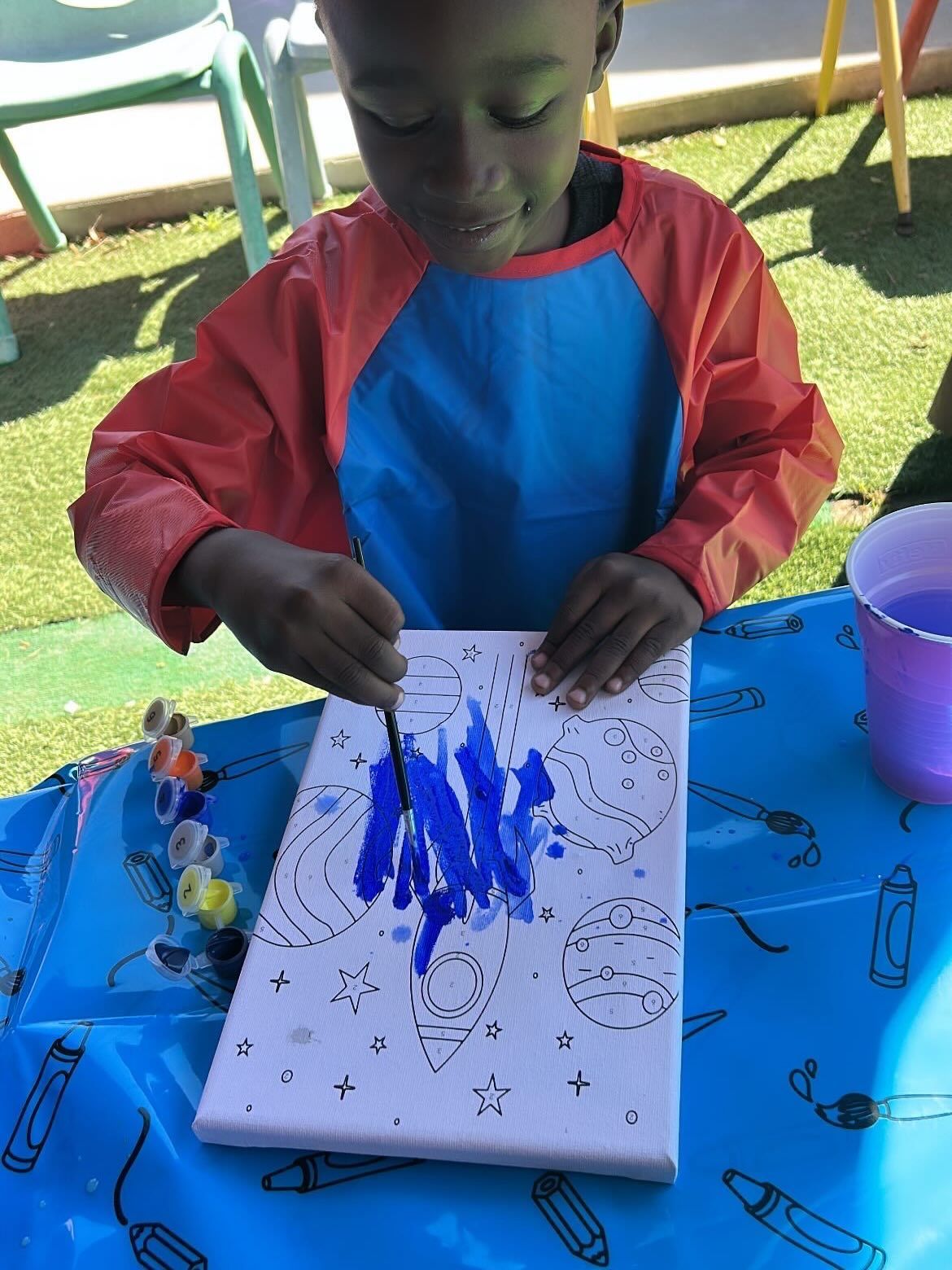 Child painting with blue paint on a canvas at an outdoor table.
